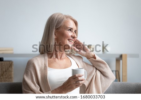 Happy dreamy middle aged woman sitting on comfortable sofa in living room with cup of black tea or coffee, looking away, Peaceful mature lady enjoying no stress calm positive pastime alone at home.