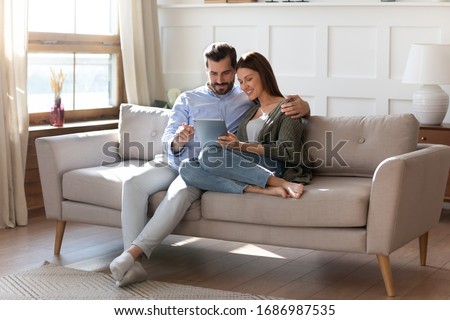 Full length affectionate young man embracing attractive wife girlfriend, looking at digital computer tablet screen, relaxing on comfortable couch. Loving couple shopping online in internet store.