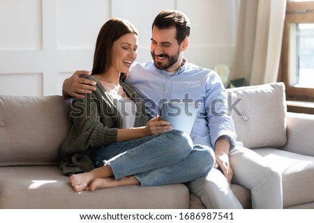 Excited young married couple resting on sofa, watching comedian funny movie on computer tablet, enjoying free weekend leisure time together. Happy family spouses cuddling, shopping online at home.