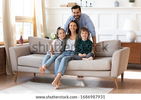 Portrait of smiling bearded father standing near couch with sitting happy wife and little children. Joyful affectionate family of four posing for photo, looking at camera, good relations concept.