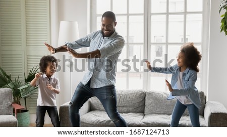 Happy African ethnicity father have fun teaches little preschool kids to dance in modern living room at home. Dad with son and daughter engaged in funny activity enjoy leisure carefree weekend concept