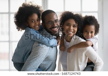 Portrait of African ethnicity parents piggy back son and daughter posing indoors. Pretty family looking at camera photo shoot in modern apartment at home, happy parenthood family services ad concept