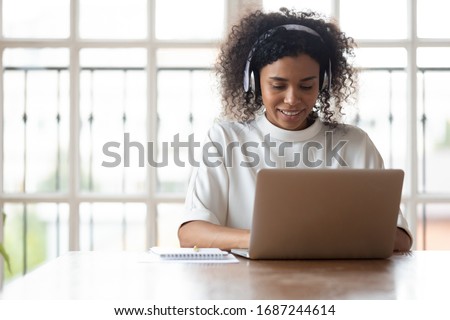 Smiling African ethnicity businesswoman wearing headphones using laptop at work, typing message during distance language studying e-learning on-line, makes video call provide support to client concept