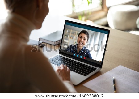 Young female sit at desk at home talk on video call with Indian colleague, woman have pleasant Webcam conference with biracial coworker, communicate online using wireless Internet connection
