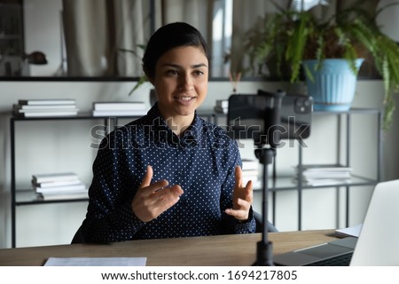 Smiling indian ethnic girl sitting in front of smartphone on stabilizer, recording self-presentation video or sharing professional skills. Happy young smart businesswoman filming educational lecture.