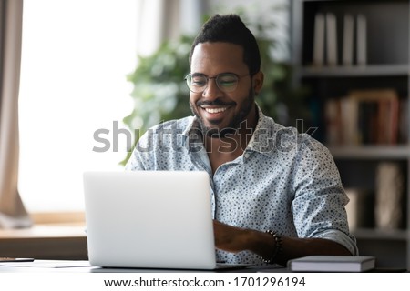 Smiling biracial man in glasses sit at desk in office browsing wireless Internet on laptop device, happy African American male worker laugh watch funny video on computer gadget, relax at work break