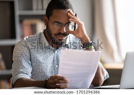 Pensive unhappy biracial man in glasses feel distressed reading bad news in paperwork letter, disappointed frustrated African American male confused by unpleasant postal correspondence or notice