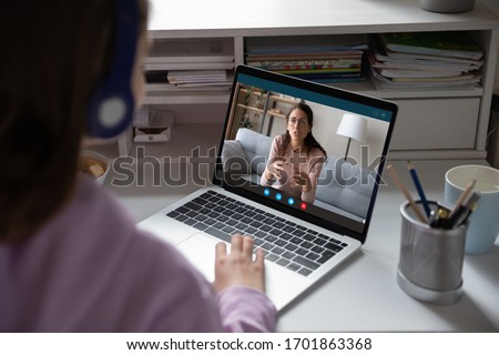 School teen girl student wears headphones distance learning with online teacher on computer screen. Web tutor gives remote class teaching teenage pupil elearning from home. Over shoulder close up view