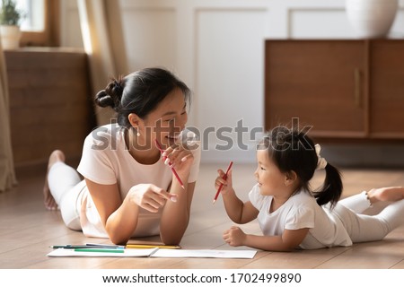 Educational pastime develop creativity skill in kid concept. Asian mother her small daughter lying on warm wooden floor in sunny cozy living room, mom teach girl paint use album and colourful pencils
