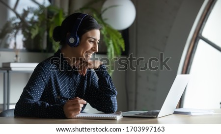 Smiling Indian female employee in wireless earphones talk on video call, have web conference with colleagues, happy ethnic woman worker in headset watch webinar on modern laptop at home