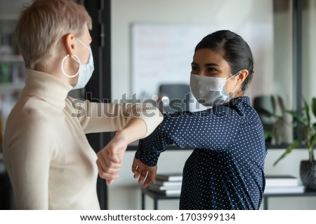 Photo of Smiling diverse female colleagues wearing protective face masks greeting bumping elbows at workplace, woman coworkers in facial covers protect from COVID-19 coronavirus in office, healthcare concept