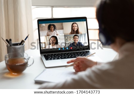 Woman sitting at desk noting writing information studying at home with multiracial students diverse ladies makes video call using video conference application, view over girl shoulder to laptop screen
