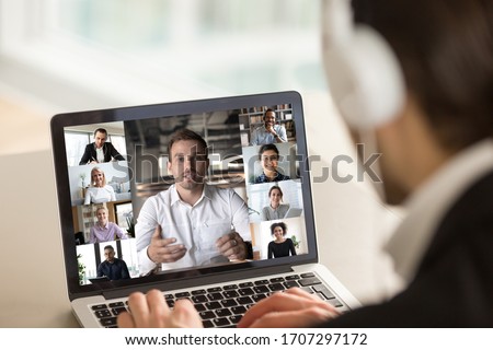 Different age and ethnicity diverse businesspeople participating at group videocall, laptop screen webcam view over man in headphones shoulder. Distant communication videoconference activity concept