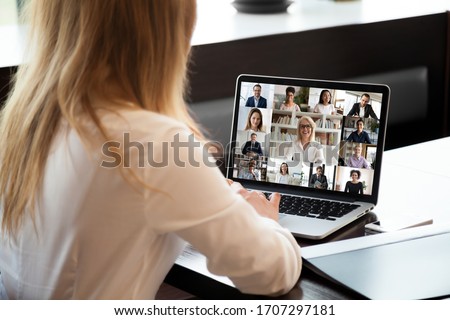 View over businesswoman shoulder sit at workplace desk looks at pc screen during group videocall with different age and ethnicity colleagues using application and webcam, distant communication concept