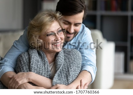 Happy senior mother spend time with grown up son, relatives people hugging at home, adult attentive millennial grateful child wrapped in a plaid or warm sweater loving mommy caring about her comfort