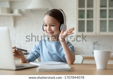 Smiling little Caucasian girl in headphones have video call distant class with teacher using laptop, happy small child wave greeting with tutor, study online on computer, homeschooling concept
