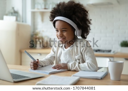 Smiling small African American girl in headphones watch video lesson on computer in kitchen, happy little biracial child in earphones have online web class using laptop at home, homeschooling concept