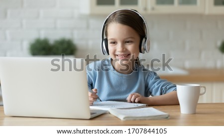 Smiling little Caucasian girl in headphones handwrite study online using laptop at home, cute happy small child in earphones take Internet web lesson or class on computer, homeschooling concept