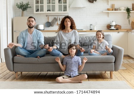Full length peaceful calm couple practicing yoga exercises with small children in studio living room. Mindful little boy sitting on floor in lotus pose while parents relaxing on sofa with sister.