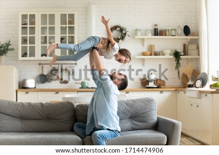 Happy young man lifting small preschool child daughter in air sitting on comfortable sofa in modern studio living room. Overjoyed kid girl having fun with handsome father, flying on hands at home.
