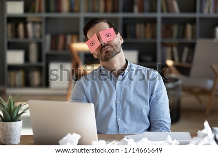 Exhausted tired businessman with painted eyes on stickers, adhesive notes on face sleeping at workplace, sitting at desk with laptop, unproductive lazy young male dozing, working on difficult project