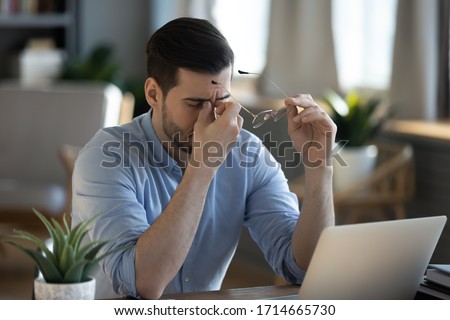 Tired exhausted businessman massaging nose bridge, taking off glasses, feeling eyestrain, sitting at work desk, young man suffering from dry eye syndrome, fatigue after long laptop use