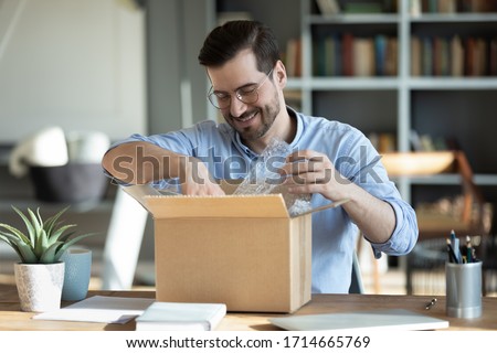 Smiling man wearing glasses unpacking awaited parcel, looking inside, sitting at work desk, satisfied happy customer opening cardboard box with online store order, good shipping delivery service 商業照片 © 