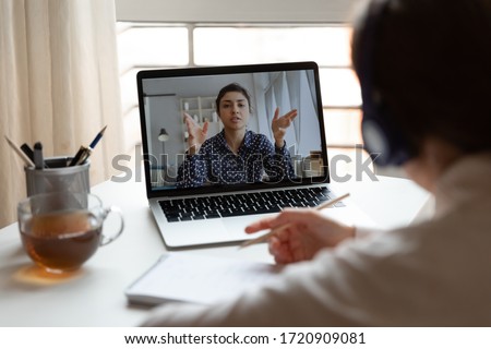 Back view of millennial girl in headphones listen watch online training or course on laptop, female employee make notes engaged in webcam conference or briefing with colleague, have web meeting at