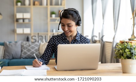 Head ahot happy indian girl student wearing headphones, watching educational lecture workshop seminar, writing notes. Smiling hindu woman studying online, sitting at desk with computer at home.