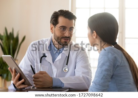 Smiling young Caucasian male doctor in white medical uniform consult female patient using tablet on consultation, modern man GP talk discuss checkup results with woman client at meeting in hospital