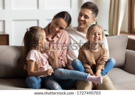 Happy young mother and father with two little daughters sitting on couch, looking at each other, family enjoying tender moment, smiling parents and preschool children having fun together