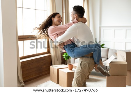 Loving husband lifting excited wife, celebrating moving day, having fun in modern living room with cardboard boxes with belongings, happy young couple purchasing new house, mortgage and relocation