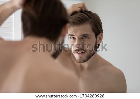 Anxious young Caucasian man look in mirror in bathroom touch check head worried about dandruff, concerned millennial male frustrated about hair loss, receding hairline, haircare, beauty concept