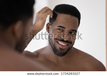 Smiling african American young man look in mirror in bathroom get ready in morning, happy biracial millennial male style hair, do daily beauty procedures treatment in bath, haircare concept