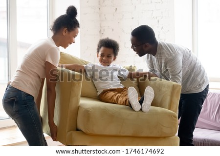 African couple carrying armchair where sit adorable small toddler son. Happy family placing delivered furniture bought in modern store, furniture shop advertisement, relocation day at new home concept