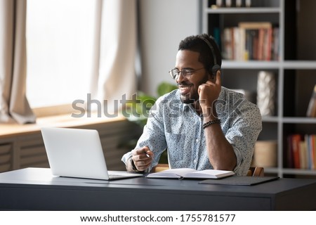 African guy student learn on-line use headphones with mic and pc holds pen takes notes in notebook sit at desk at home. Businessman wear headset talk by conference call do video chat working at office