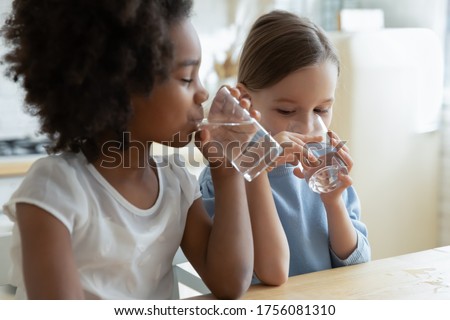 Two multi racial little girls sit at table in kitchen feels thirsty drink clean still natural or mineral water close up image. Healthy life habit of kids, health benefit dehydration prevention concept Foto d'archivio © 