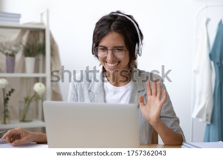 Smiling young Caucasian woman in headset wave greet talking on webcam virtual conversation on laptop, happy female in wireless headphones speak on video call on computer, consult client online