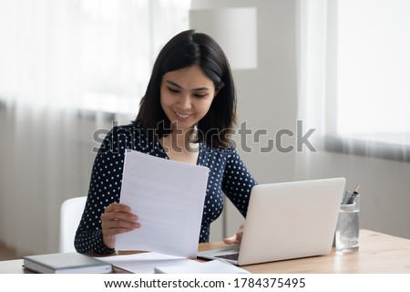Happy millennial Asian girl sit at desk work on laptop consider read good news in post paper correspondence or letter document, smiling Vietnamese young woman study on computer prepare report