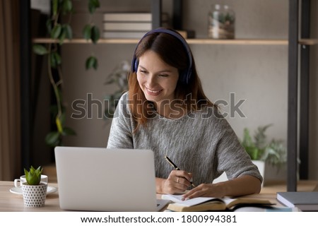 Smiling millennial Caucasian girl in headphones sit at desk look at laptop screen study online, happy smart young woman in earphones take web course or training on computer, distant education concept