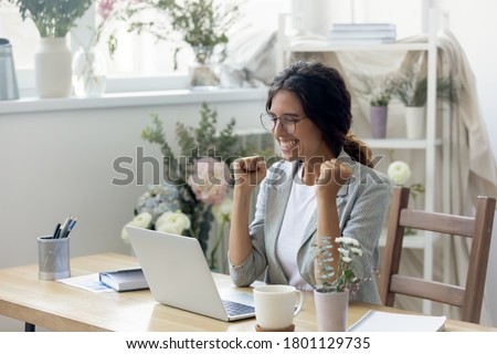 Euphoric young creative designer looking at laptop screen, celebrating receiving good news at modern showroom. Emotional happy businesswoman in glasses getting bank loan approval or making first sake.