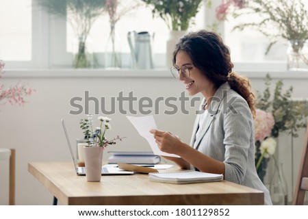 Side view pleasant smiling young businesswoman creative designer in eyeglasses reading paper letter correspondence with good news or banking notification, sitting at desk in modern showroom office.