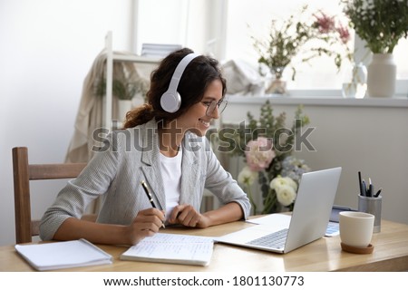 Happy young businesswoman wearing earphones, enjoying studying online, writing notes. Smiling creative designer decorator in eyeglasses looking at computer screen, improving professional skills.