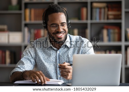Smiling African American man wearing headphones looking at laptop screen, motivated student writing notes during online lesson, watching webinar, learning language online, sitting at work desk