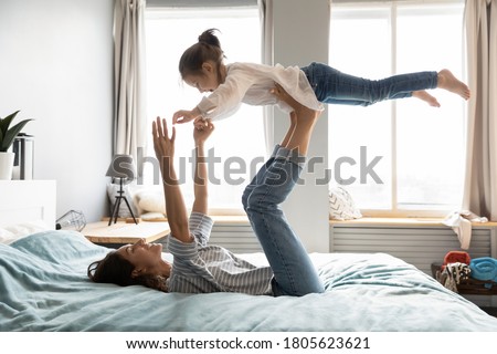 Full length strong young smiling mommy lying on comfortable bed, holding small kid daughter on straight legs. Happy little child girl flying in air, practicing yoga activity with mother in bedroom.