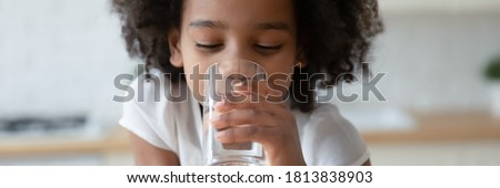Thirsty funny small african ethnicity cute kid girl drinking fresh pure water, refreshing during day or enjoying morning healthcare routine, horizontal photo banner for website header design.
