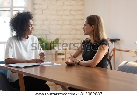 Smiling African American young businesswoman talk negotiate with female Caucasian job candidate at office meeting, happy biracial boss discuss ideas with business partner or employee at interview