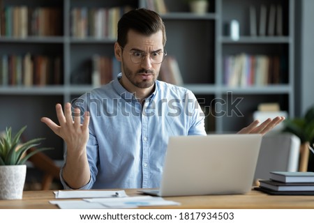 Unhappy young Caucasian male worker in glasses look at laptop screen shocked by gadget breakdown or operational problems. Frustrated man confused surprised by unexpected error on computer device.