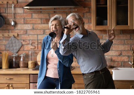 Happy senior husband and wife have fun sing in kitchen appliances cooking together at home. Overjoyed mature grey-haired Caucasian couple feel energetic active enjoy family retirement weekend.
