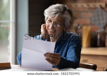 Happy mature 60s woman in glasses sit at table manage paperwork read postal letter correspondence. Smiling elderly Caucasian female in spectacles consider post document or bank paper notice.